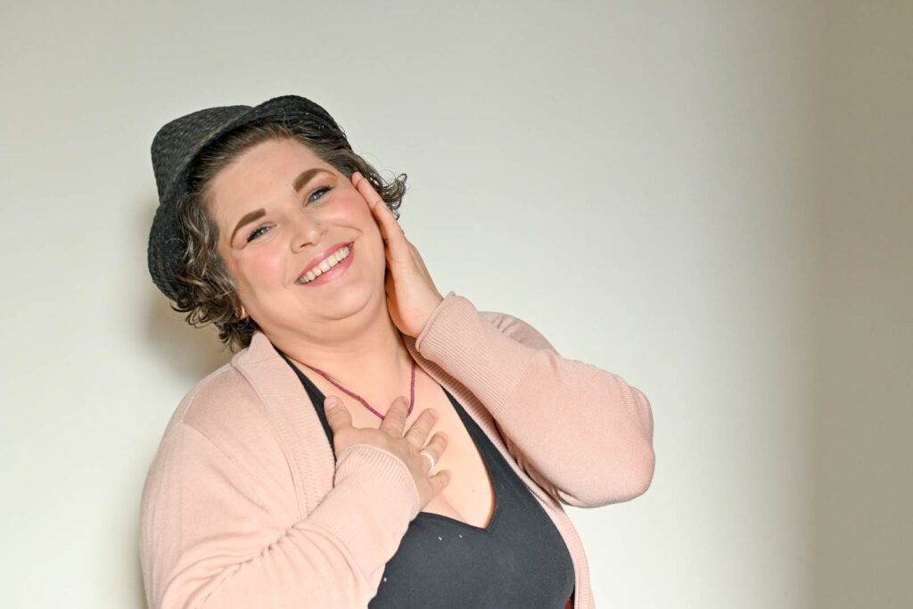 Sarah is standing against a white wall. Her short wavy hair is underneath a black hat. She is smiling, and has one hand on her cheek and one on her heart. She is wearing a light pink cardigan and a black shirt underneath with a red necklace.