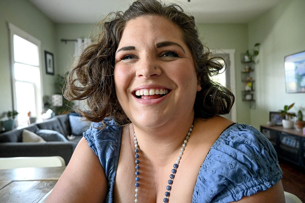 Sarah Wallace West Kootenay Postpartum Doula close up from chest up. She's smiling and wearing a blue short sleeve top as well as a blue beaded necklace that hangs down out of the photo. Her hair is at her chin and wavy, pulled back at the top but loose everywhere else.