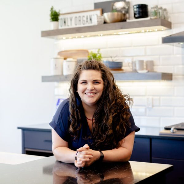 Sarah leans forward on a counter in a beautiful kitchen. She is wearing a blue dress and her long, wavy hair is loose. She is smiling and her hands are clasped together.